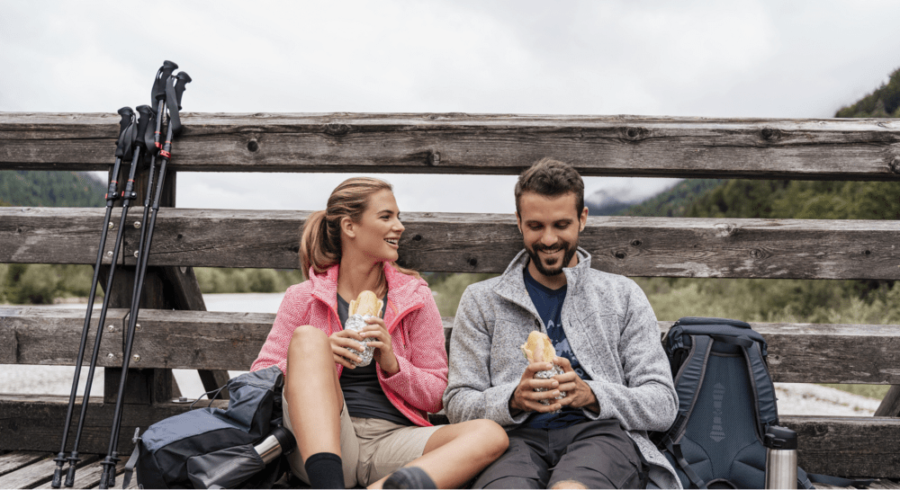 Ungt par på tur sitter på bakken inntil et gjerde mens de spiser nisten sin.
